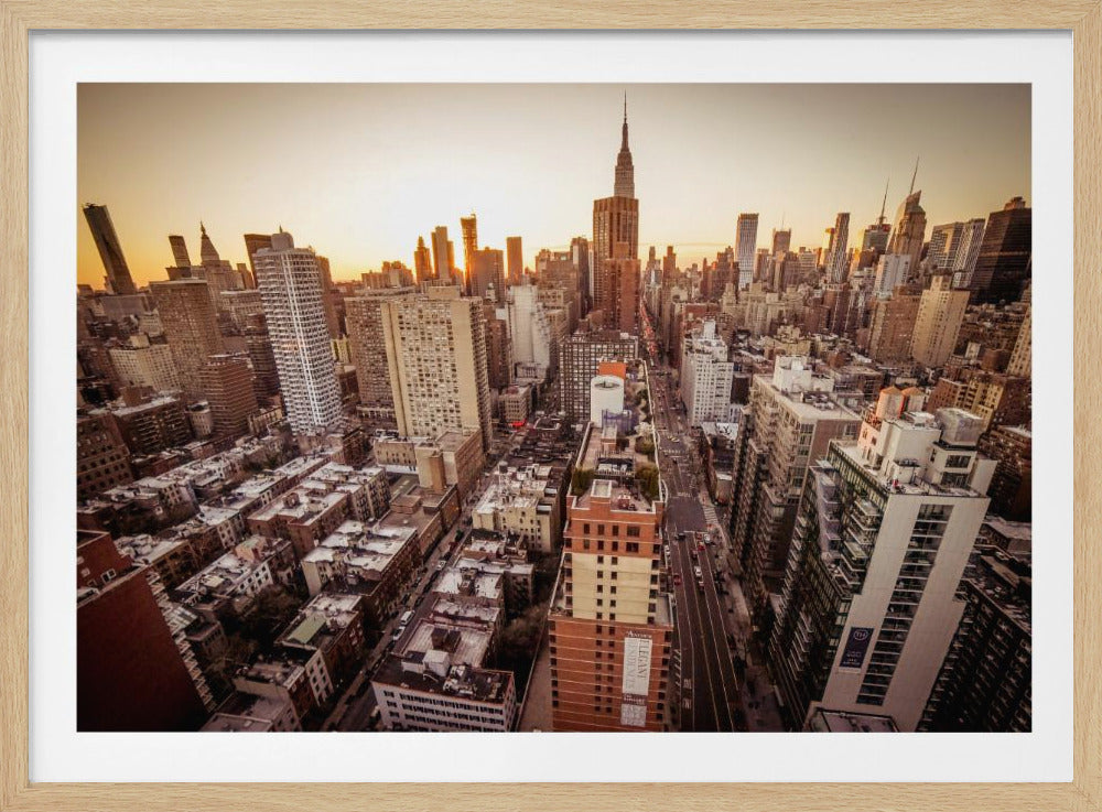 An aerial photograph of the New York City skyline at sunset, with the Empire State Building prominent against the glowing sky. The warm light of the setting sun bathes the dense urban landscape in a golden hue. The entire scene is enclosed in a silver, brushed metal frame. Print