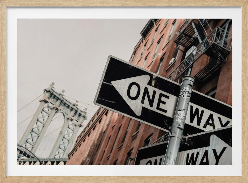 A low-angle shot of a black and white 'ONE WAY' street sign in the foreground, with a red brick building and the Manhattan Bridge visible in the background under an overcast sky, all enclosed in a silver frame. Artwork
