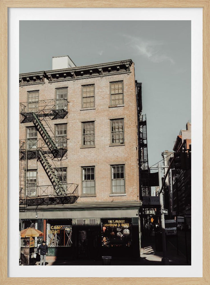 A low-angle, desaturated photograph of a classic brown brick building in a city, featuring multiple windows and a black metal fire escape zigzagging up the side. The sky is a pale, hazy blue, and the ground floor has dark storefronts. Decor