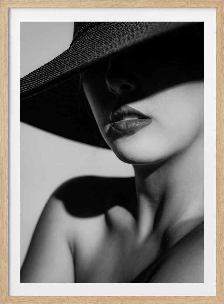 A dramatic black and white close-up photograph of a woman wearing a large, dark, wide-brimmed hat. The hat casts a deep shadow over the upper half of her face, obscuring her eyes and creating a sense of mystery. Only her nose, full lips, chin, neck, and shoulder are visible, highlighted by a strong light source. Decor
