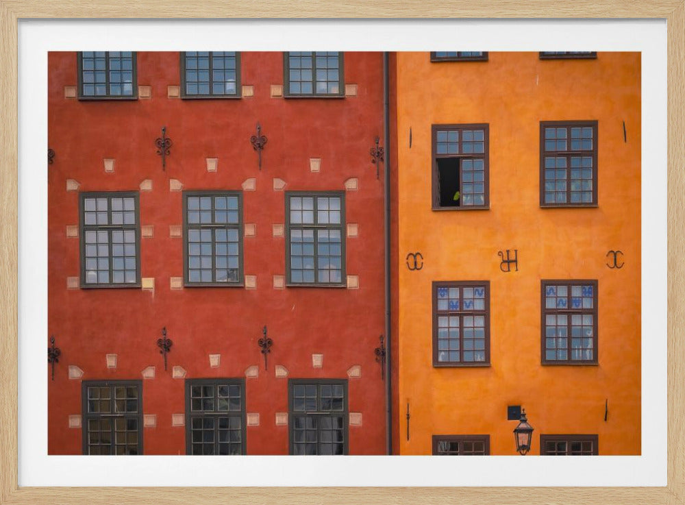 A framed close-up photograph of two adjacent, colorful building facades. The building on the left is a deep, rustic red, while the one on the right is a vibrant orange. Both feature numerous multi-paned windows with dark frames. Artwork