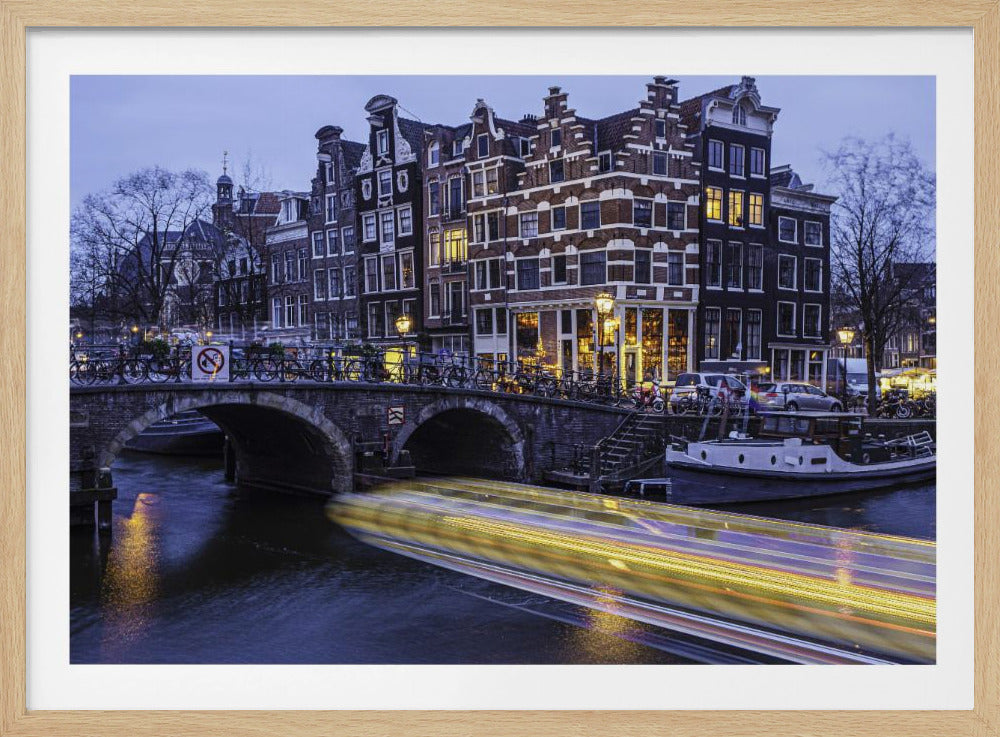 A long-exposure photograph of an Amsterdam canal at dusk, featuring a bright yellow light trail from a passing boat. In the background, illuminated historic canal houses line the water, and an arched stone bridge covered in bicycles crosses the canal. The entire image is presented in a silver frame. Print