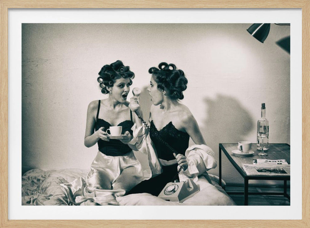 A vintage black and white photo of two women with their hair in curlers, sitting on a bed and sharing a phone call on a rotary phone. Both women have shocked expressions, suggesting they are listening to exciting gossip. One holds a coffee cup, and a bedside table with a bottle is nearby. Poster