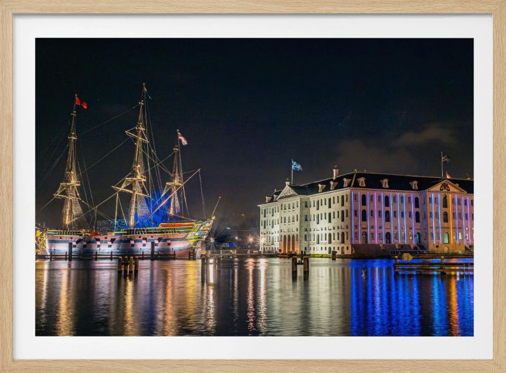A vibrant nighttime photograph of the National Maritime Museum and a replica of the East Indiaman Amsterdam ship in a harbor. Both are illuminated with colorful lights that cast beautiful reflections on the dark water. Wall Art
