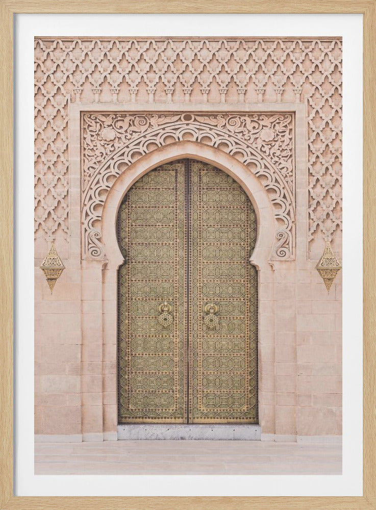 A framed photograph of an ornate golden double door set within a carved stone horseshoe arch. The surrounding facade is made of light beige stone with intricate geometric and floral carvings, characteristic of Moroccan architecture. Two decorative lanterns hang on either side of the archway. Poster