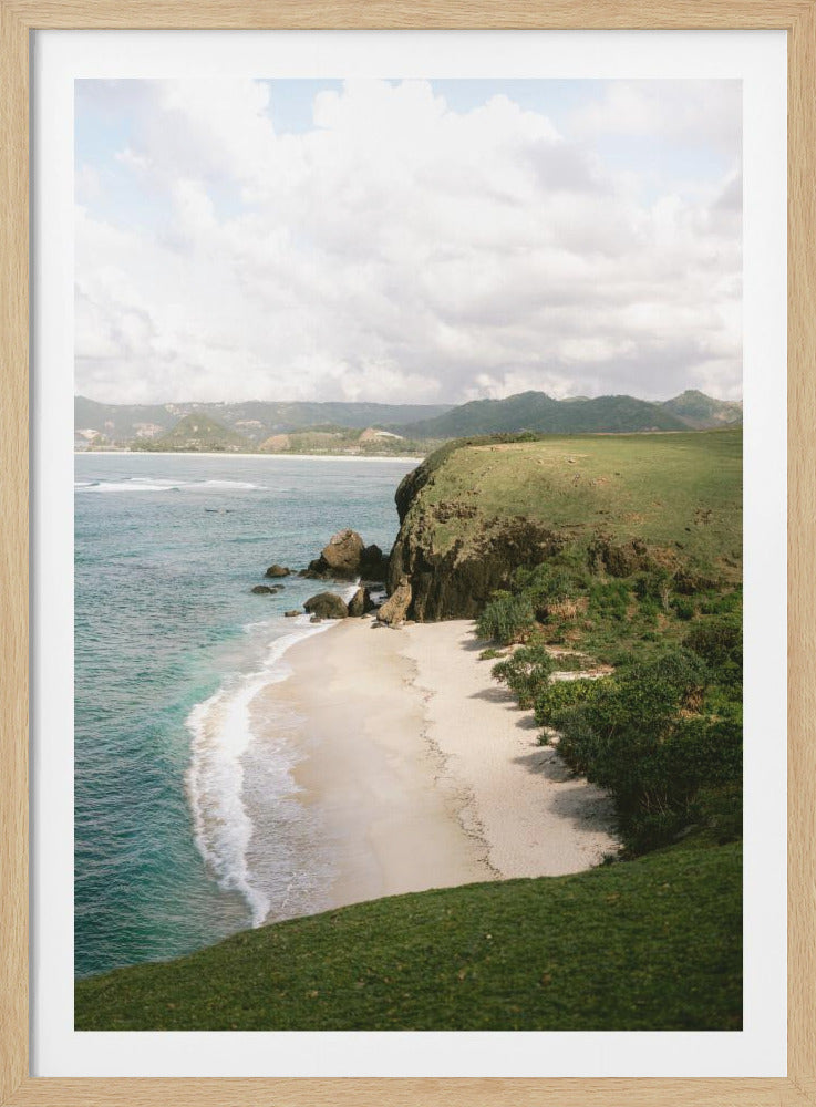 A high-angle photograph of a secluded tropical beach, with turquoise waves gently washing onto the white sand. A lush green cliff covered in grass and small trees frames the cove, with distant rolling hills visible under a soft, cloudy sky. Decor