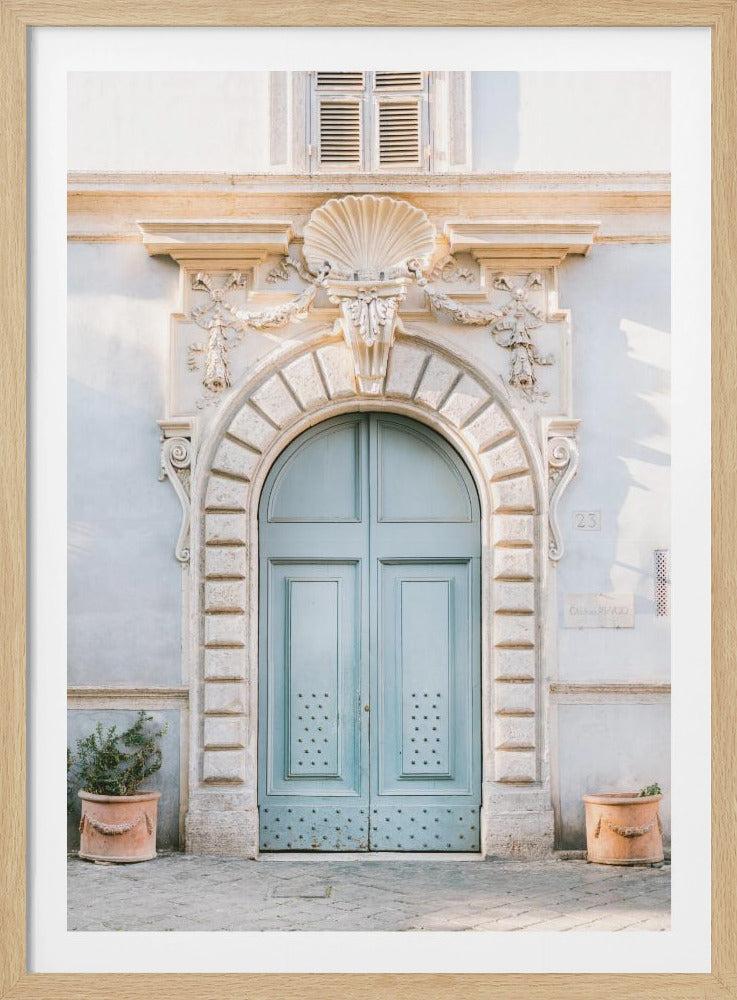 A vertical photograph of an elegant, light blue arched double door, set within an ornate, cream-colored stone entryway with a seashell motif at its apex. The building's wall is a pale off-white, and two terracotta planters with small green plants sit on the stone ground on either side of the entrance. Poster
