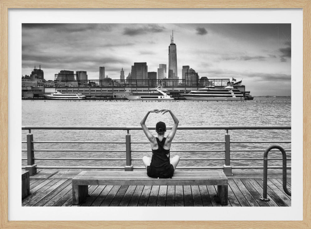 A framed black and white photograph showing the back of a woman sitting on a wooden bench on a pier. She is in a yoga pose with her arms raised, looking out at the Lower Manhattan skyline, including the Freedom Tower, across the water. Decor