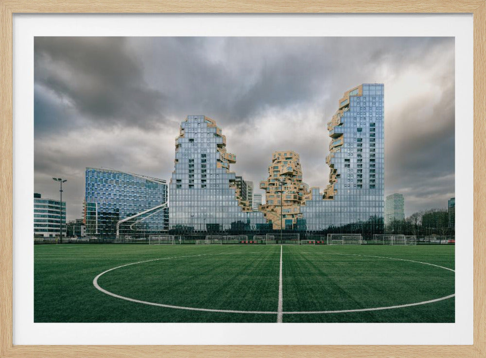 A wide-angle view from the center of a lush green soccer field looking towards a striking modern architectural complex with uniquely shaped glass and tan buildings under a dramatic, cloudy sky. Decor
