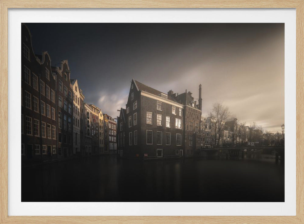 A moody, atmospheric photograph of historic canal houses in Amsterdam, viewed from the water. The scene is dark, with a dramatic, cloudy sky and a soft glow on the horizon, while some windows in the dark brick buildings are lit from within. The image is bordered by a silver frame. Wall Art