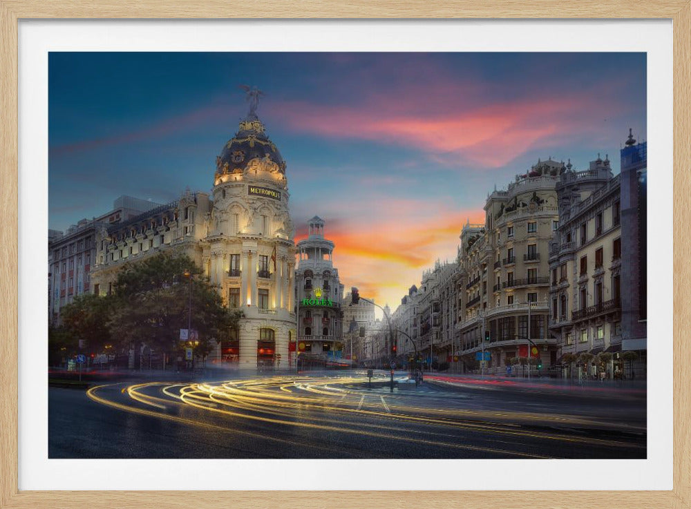 A long-exposure photograph of the Metropolis Building in Madrid at sunset, with glowing light trails from traffic swirling on the streets below a vibrant pink, orange, and blue sky. Artwork