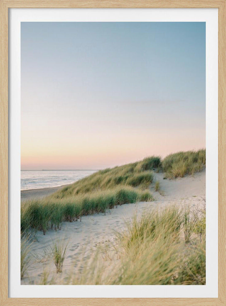 A tranquil beach scene at sunrise or sunset, featuring rolling sand dunes covered with lush green beach grass. The calm ocean is visible on the left under a soft pastel sky that gradients from pale pink to light blue. The photograph has a serene and soft-focus quality, and is displayed within a thin black frame. Wall Art