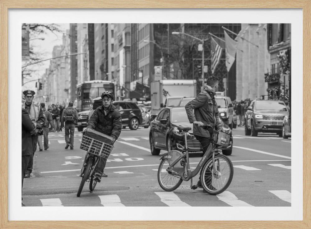 A black and white street photograph of a busy city intersection, framed in silver. In the foreground, two men are crossing with their bicycles amidst traffic, with one cyclist looking directly at the camera. Tall buildings and other pedestrians line the background, capturing a moment of everyday city life. Artwork