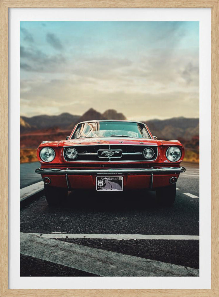 A low-angle, head-on shot of a vintage red Ford Mustang parked on an asphalt road. The iconic silver horse emblem is visible on the grille. In the background, a desert mountain range is blurred under a cloudy, muted sky. Poster