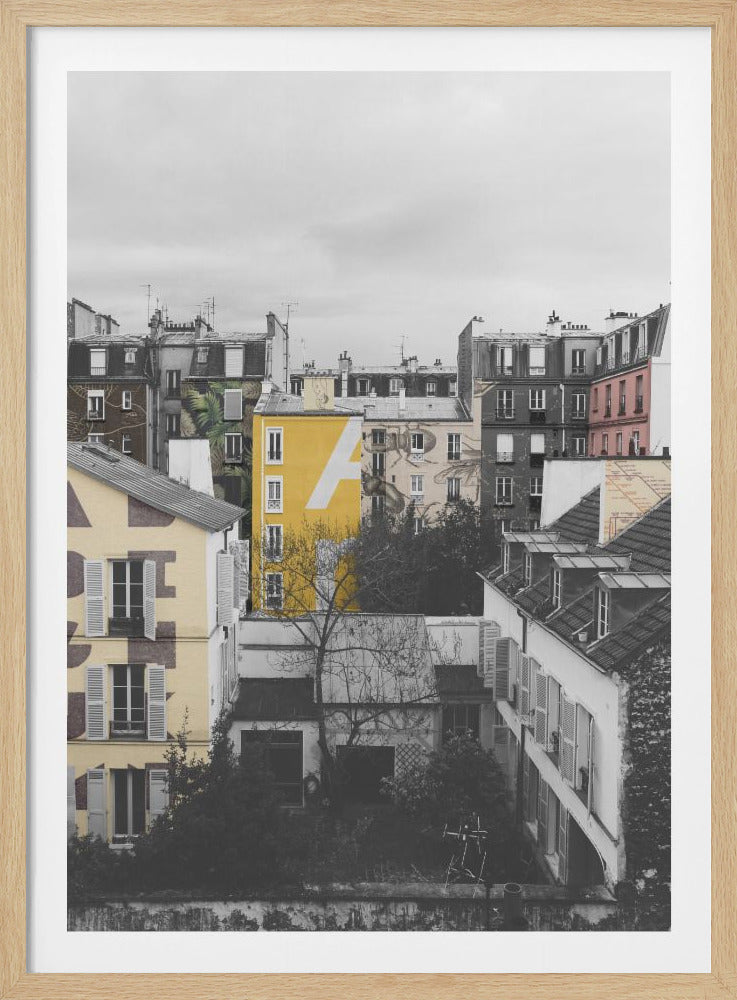 A framed, vertical photograph with selective color, showing a dense cluster of Parisian-style buildings under an overcast sky. Most of the image is in black and white, but one building is a vibrant yellow with a large white 'A' painted on it, creating a striking focal point. Wall Art