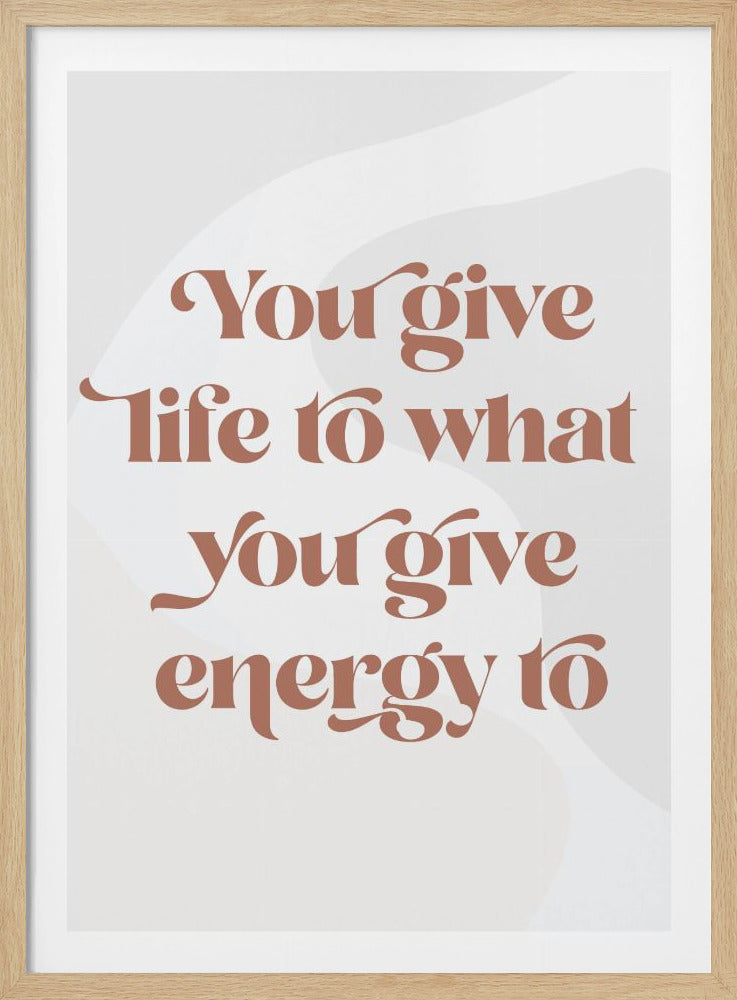 A framed motivational poster featuring the quote 'You give life to what you give energy to'. The text is printed in a dusty rose color using a stylish, retro serif font on a light gray background with subtle wavy shapes. Poster