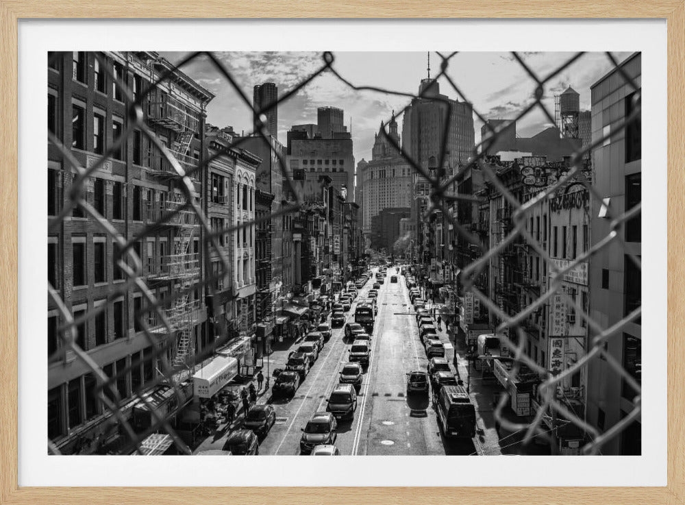 A high-angle, black and white photograph of a bustling New York City street viewed through a chain-link fence. The fence is in the foreground, framing the scene of traffic-filled streets, historic buildings with fire escapes, and skyscrapers in the distance under a cloudy sky. Wall Art