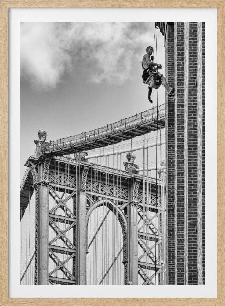 A black and white photograph capturing a window washer rappelling down the side of a tall brick building. In the background, the intricate steel structure of the Manhattan Bridge stands against a partly cloudy sky, creating a powerful urban scene. Decor