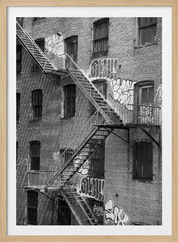 A black and white photograph of a brick apartment building with a prominent metal fire escape zigzagging across its facade. The walls are adorned with various styles of graffiti, and the windows are covered with bars, creating a gritty, urban scene. Decor