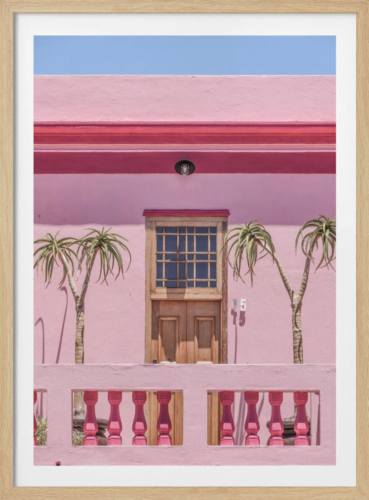 A brightly colored pink building stands under a clear blue sky, framed in black. A central wooden door with a paned window is flanked by two slender, palm-like trees. In the foreground, a pink balcony wall features fuchsia-colored balusters. Decor
