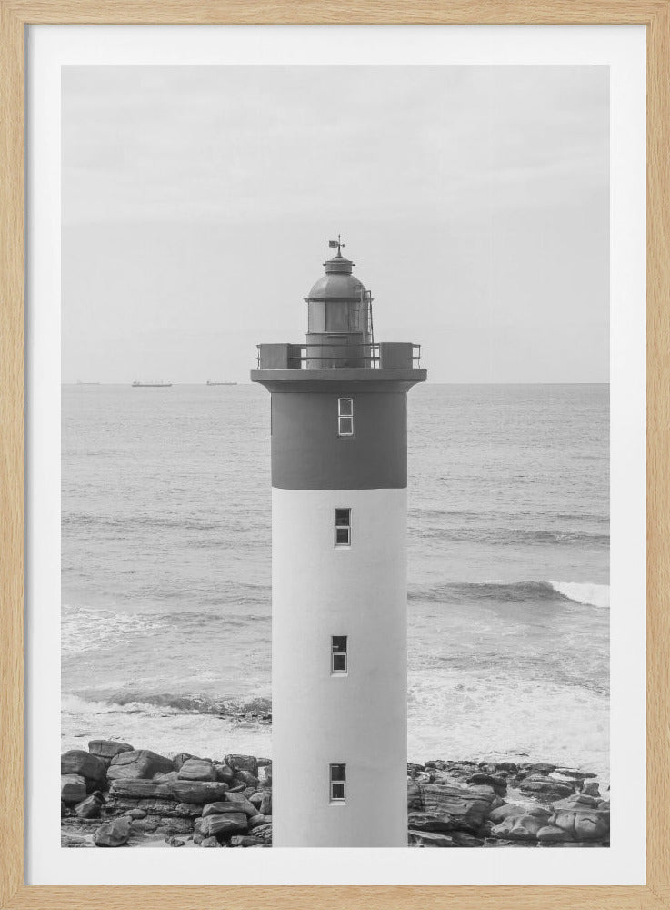 A framed, black and white photograph of a tall, cylindrical lighthouse standing on a rocky shore. The lighthouse is dark grey on its upper section and white on the lower, with the ocean and a grey sky in the background. Print