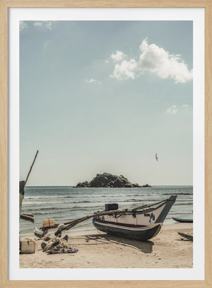 A framed photograph of a traditional outrigger fishing boat resting on a sandy beach. In the background, the ocean stretches out to a small rocky island under a pale blue sky with a few white clouds. The scene has a calm and slightly vintage feel. Decor