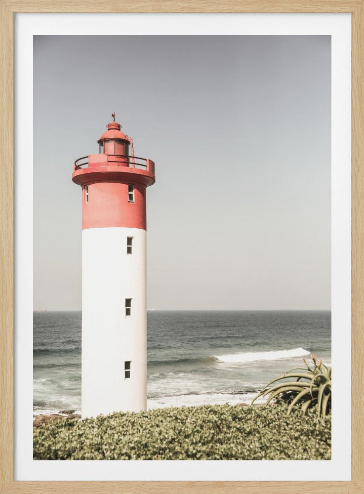 A photograph of a red and white lighthouse on the coast. The top section of the lighthouse is red, and the bottom is white. It stands on a green, bushy cliffside with the ocean and a pale, hazy sky in the background. Artwork