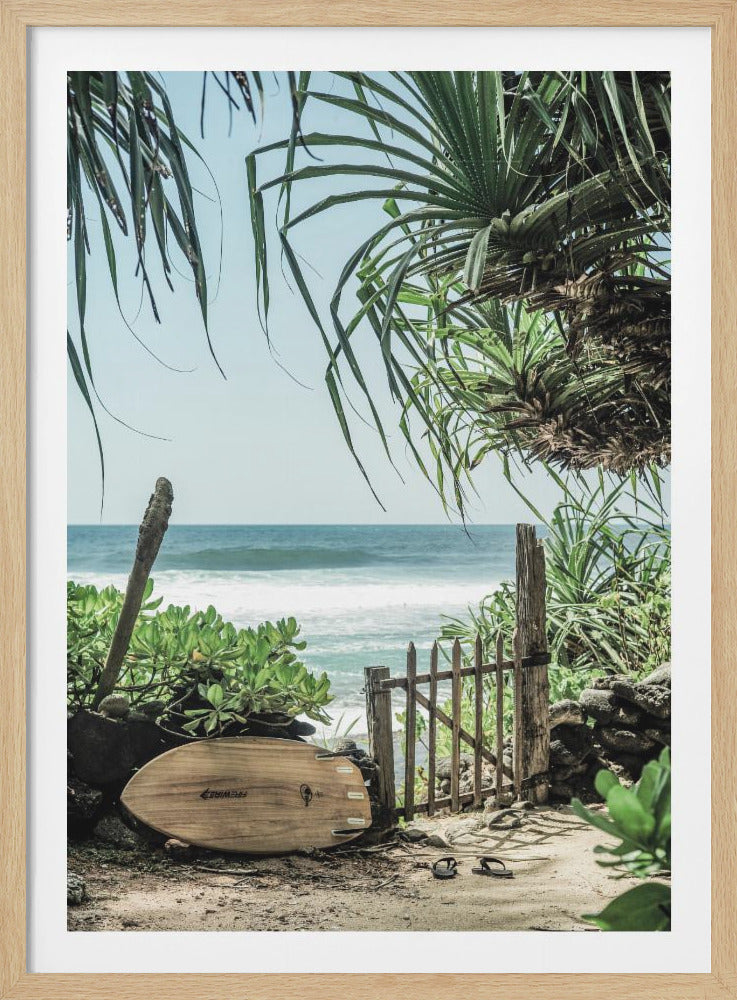 A serene view of the ocean from a sandy path framed by lush green tropical plants and palm fronds. A light wood-colored surfboard rests against rocks, next to a rustic wooden gate that opens towards the beach. A pair of flip-flops sits on the sand, and gentle waves roll onto the shore under a pale blue sky. Decor