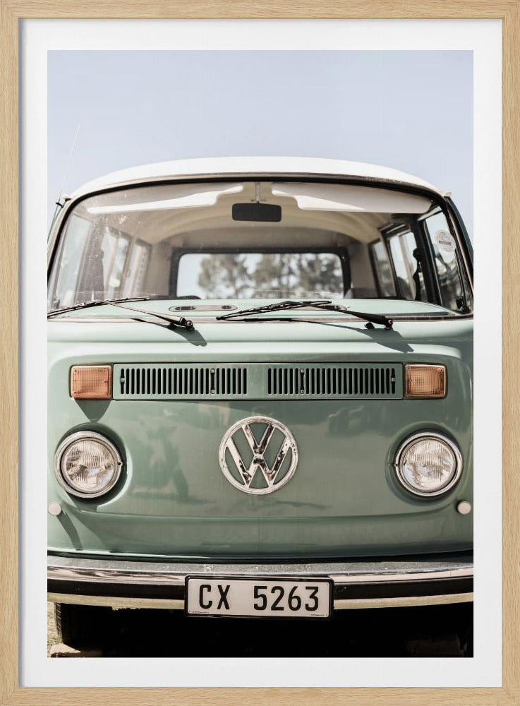 A close-up front view of a vintage sage green Volkswagen bus with a white roof. The iconic VW logo is on the front, with round headlights and a European license plate, set against a pale blue sky. Print