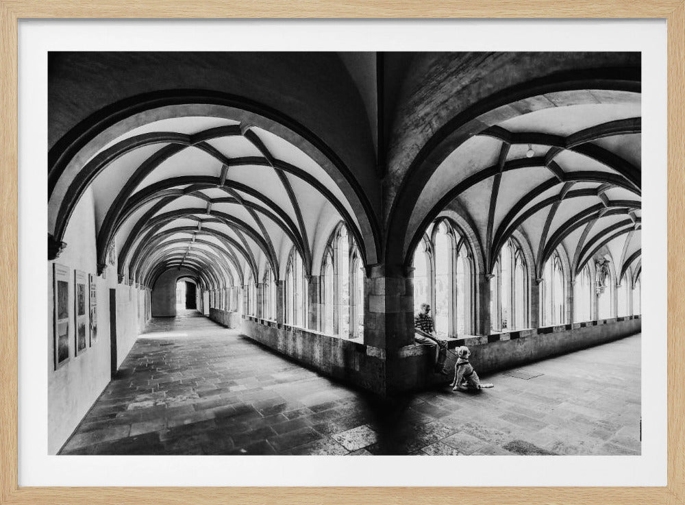 A black and white photograph of a person and their dog sitting in the corner of a Gothic cloister. The stone hallway features a dramatic vaulted ceiling and a long row of arched windows, creating a powerful sense of perspective and light. Decor