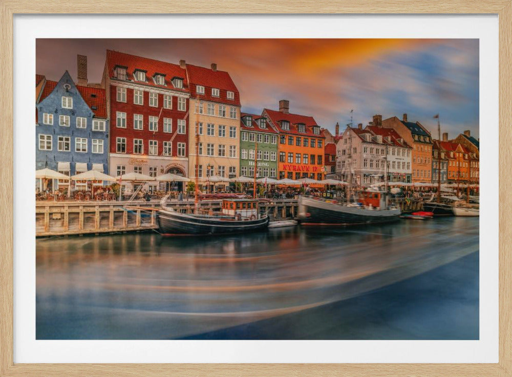 A long-exposure photograph of the colorful Nyhavn harbor in Copenhagen at sunset. Historic, brightly colored buildings line the canal, where two traditional wooden boats are moored. The sky is a dramatic blend of orange and blue, and the water is blurred, showing streaks of reflected light. Decor