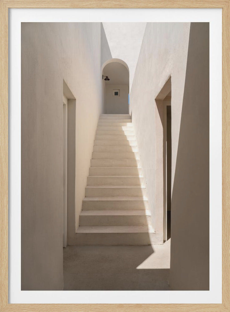 A minimalist architectural photograph of a narrow corridor with beige walls leading to a flight of stairs. Sunlight casts strong, sharp shadows, and the stairs ascend towards a distant arched doorway. Wall Art
