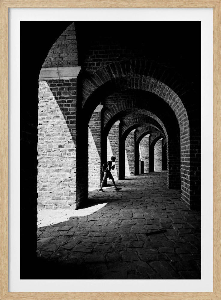 A dramatic black and white photograph of a person walking through a long brick arcade. Sunlight creates strong, contrasting shadows, highlighting the repeating arches that recede into the darkness, creating a powerful sense of depth and perspective. Decor
