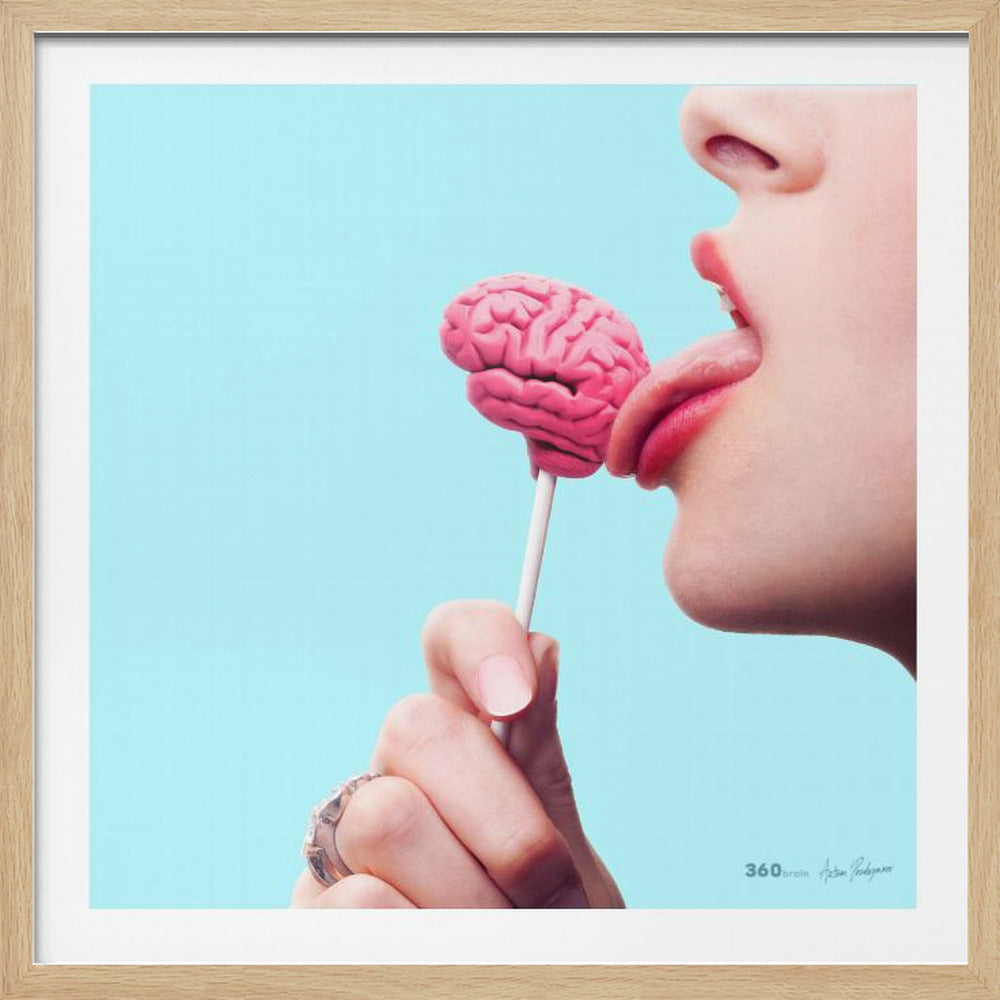 A conceptual art piece in a light wood frame, featuring a close-up of a person licking a bright pink lollipop shaped like a human brain against a solid light blue background. Artwork