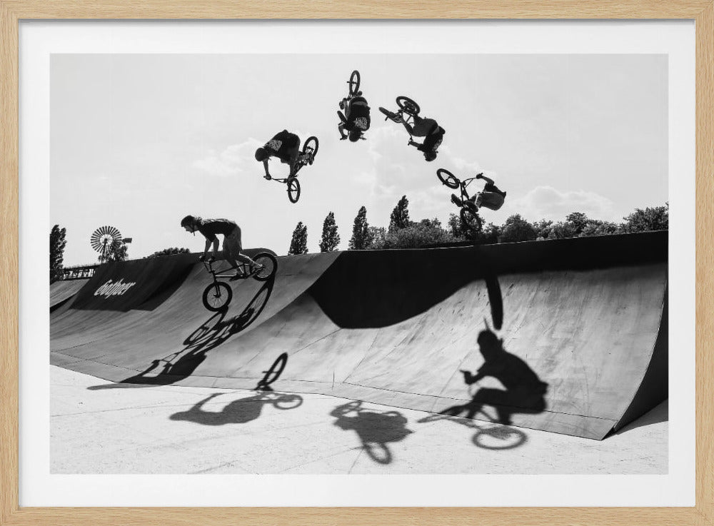 A dynamic black and white action shot showing the progression of a BMX rider doing a backflip off a large ramp. Multiple exposures capture the rider at different points in the air, creating an arc of motion against a bright sky. Strong shadows are cast on the ramp below. Print