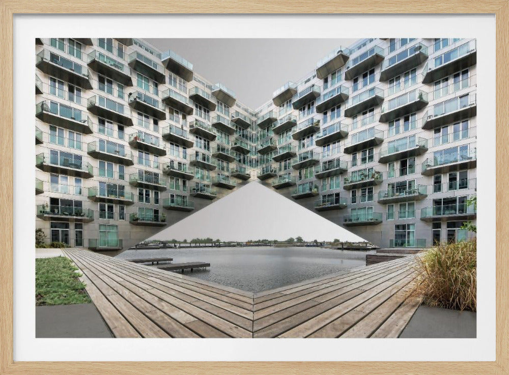 A symmetrical, mirrored photograph of a modern V-shaped apartment complex with numerous glass balconies, overlooking a wooden deck and a calm body of water under a grey sky. The image is presented within a silver picture frame. Artwork