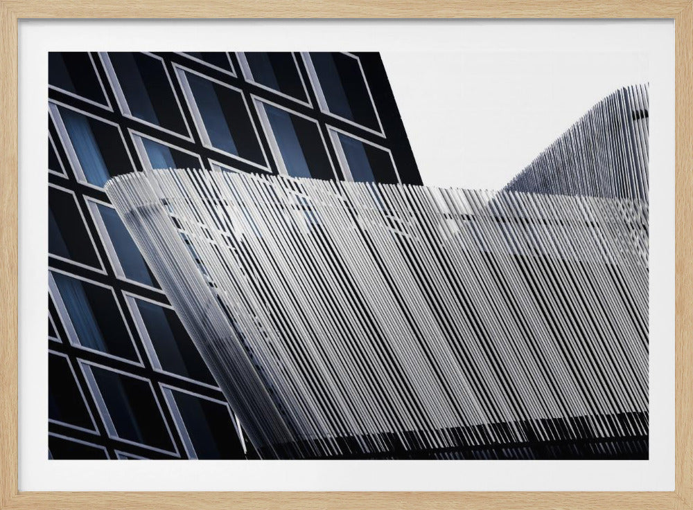 An abstract architectural photograph, framed in silver, showing a dynamic contrast between two modern building facades. In the background, a dark building features a grid of diamond-shaped windows with white frames, while a structure in the foreground creates a wave of fine, parallel white vertical lines against the sky and the other building. Print