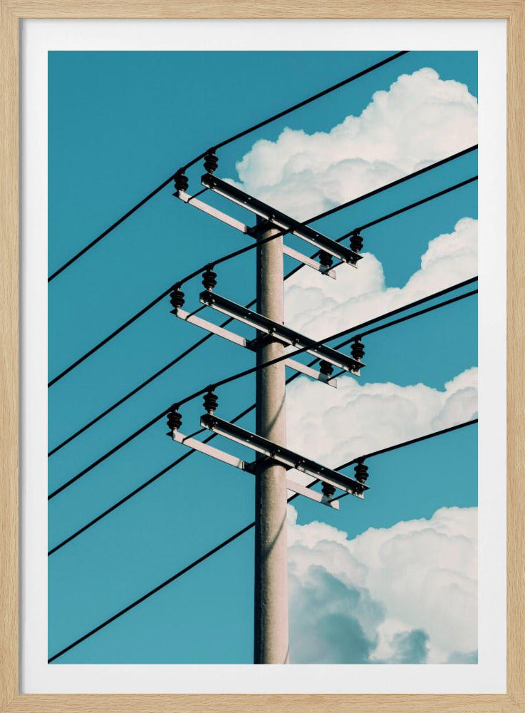 A low-angle photograph of a concrete utility pole with black power lines stretching diagonally across the frame against a vibrant blue sky with fluffy white clouds. Decor