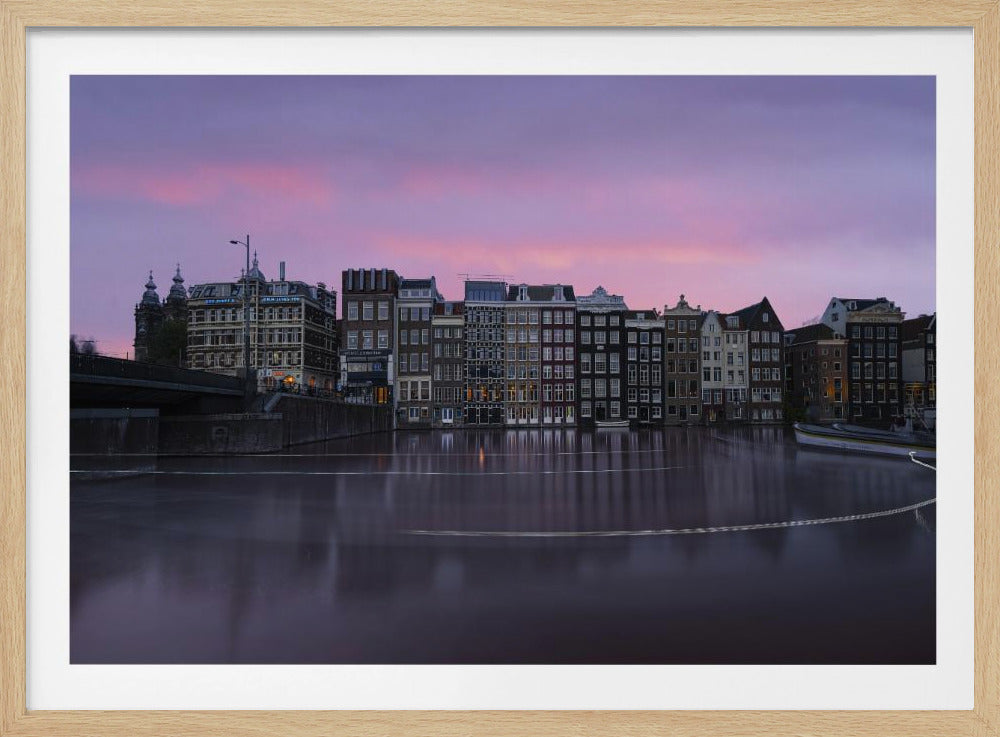 A framed, long-exposure photograph of historic Amsterdam canal houses lining the water at dusk. A vibrant purple and pink sky is reflected in the smooth surface of the canal, where faint light trails from a boat are visible. Wall Art