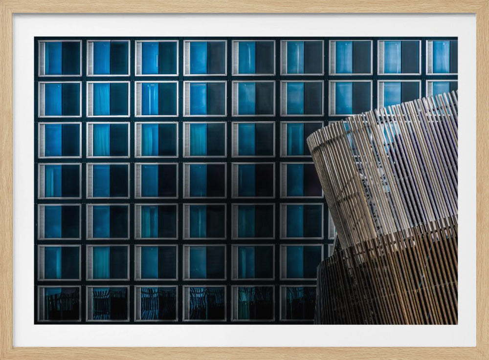 An architectural abstract photograph featuring a modern building facade with a grid of blue and black square windows, contrasted by a curved, slatted structure in the foreground, all enclosed in a silver frame. Wall Art