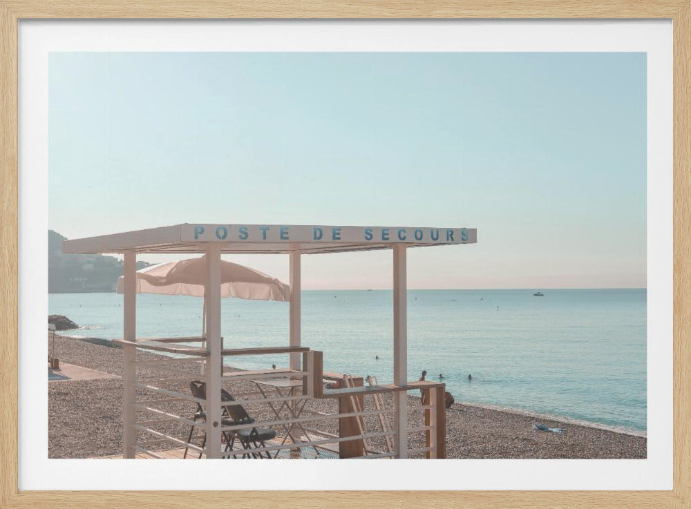 A framed photograph of a serene white lifeguard station on a pebble beach, with a sign reading 'POSTE DE SECOURS'. The calm, light blue ocean stretches to the horizon under a hazy, pale sky, creating a peaceful, pastel-toned scene. Wall Art
