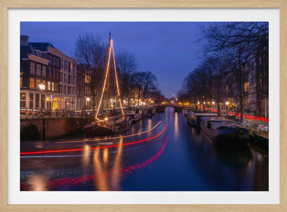 A long-exposure photograph of an Amsterdam canal at dusk. The scene is bathed in a deep blue light, with warm yellow and orange lights from street lamps and buildings reflecting on the water. A sailboat is decorated with glowing string lights, and red and white light trails from a passing boat create streaks across the canal. Houseboats are moored along the sides, and a bridge is visible in the distance. Poster
