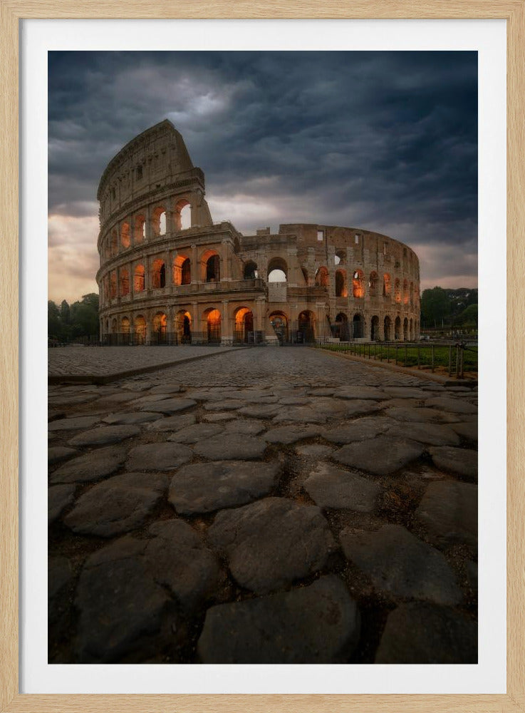 A low-angle view of the Roman Colosseum at dusk, with its arched windows glowing with warm orange light under a dramatic, stormy sky. An ancient cobblestone road leads towards the iconic amphitheater. Print