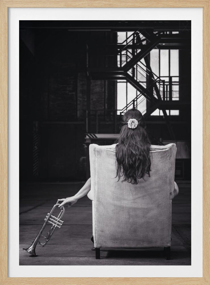 A black and white photograph of a woman seen from behind, sitting in a large, light-colored corduroy armchair. She has long, dark hair with a white flower pinned in it. Her left hand holds a trumpet, resting it on the dark floor of what appears to be an industrial warehouse with a metal staircase in the background. Wall Art