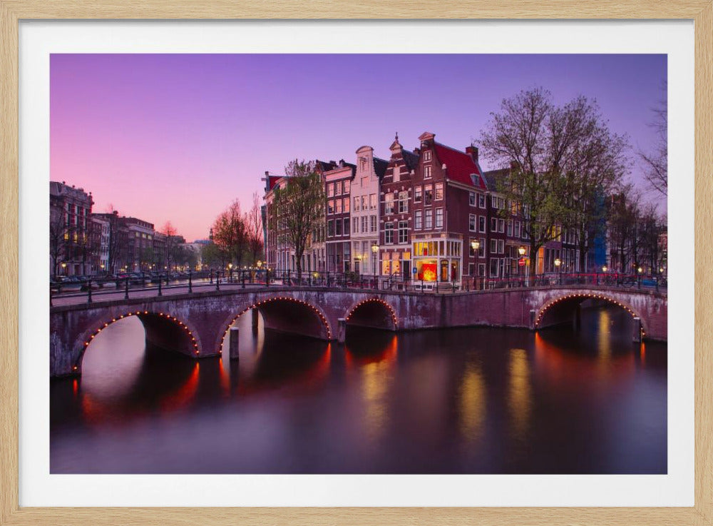 A scenic view of an Amsterdam canal at twilight, with historic brick buildings and illuminated arched bridges reflected in the calm water under a vibrant pink and purple sky. Poster