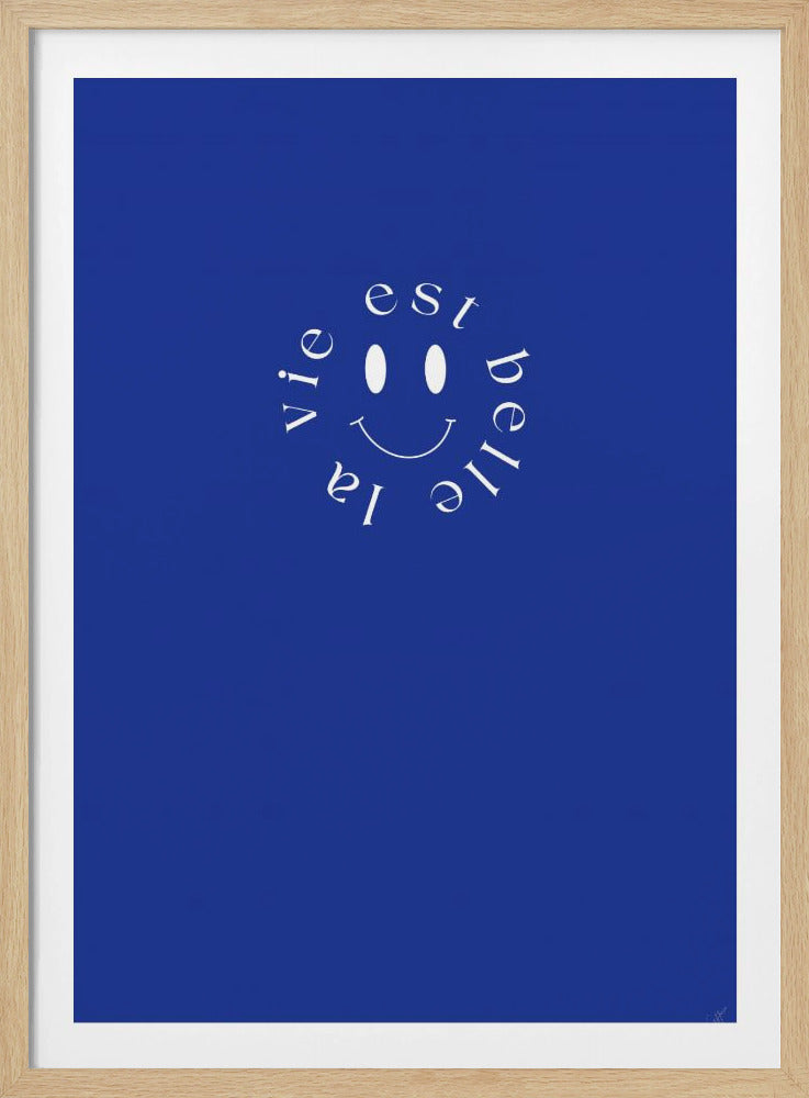 A minimalist poster with a vibrant royal blue background. In the upper center, a white smiley face is surrounded by the French words 'la vie est belle' (life is beautiful) arranged in a circle. The entire piece is enclosed in a simple black frame. Wall Art