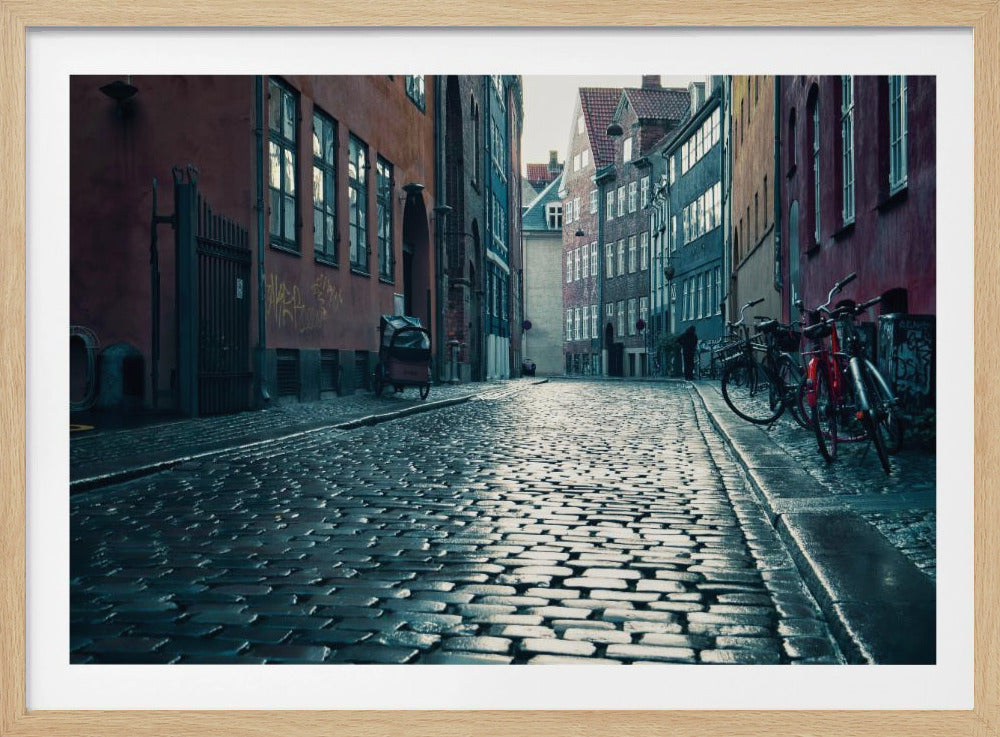 A low-angle shot of a wet, reflective cobblestone street in a narrow European alley. The street is lined with historic, colorful buildings in shades of red, orange, and blue under an overcast sky. Bicycles are parked along the sidewalk on the right, adding to the quiet, moody city scene. Poster