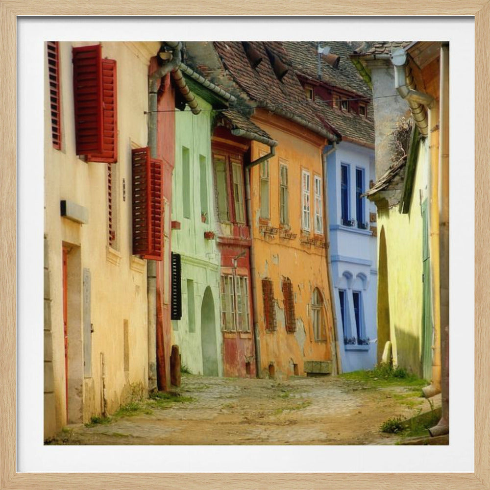 A narrow cobblestone street winds through a historic European town, flanked by colorful, weathered buildings in shades of cream, green, orange, and blue with red shutters, all under aged tile roofs and enclosed in a light wood frame. Artwork