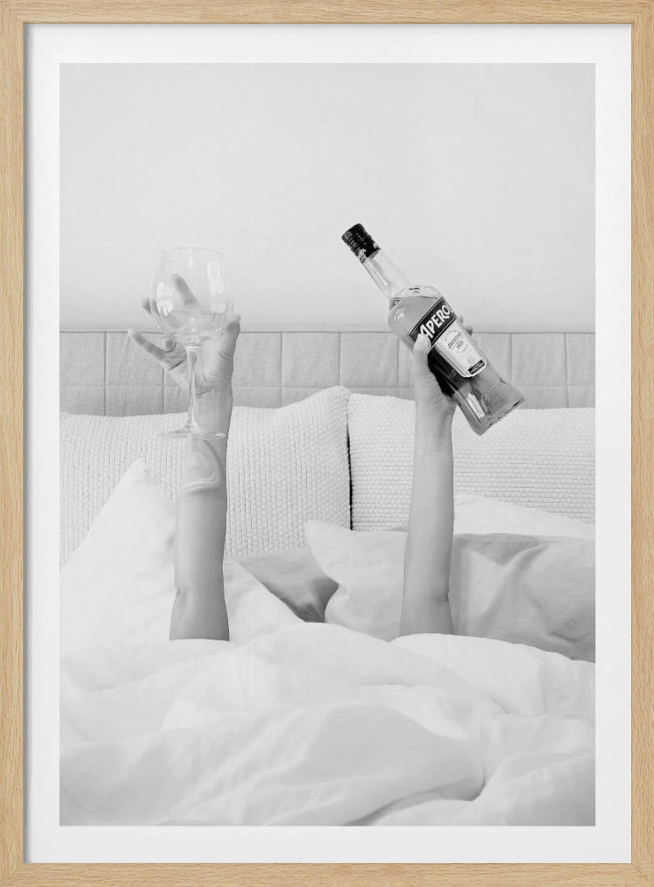 A black and white photograph of a person under white covers in bed, with only their arms visible. One hand holds up a bottle of Aperol, and the other holds up an empty wine glass. Decor