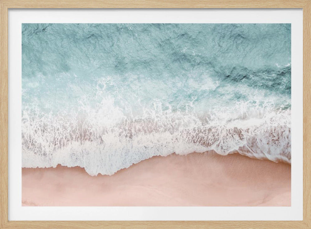 An aerial, top-down view of a turquoise ocean wave with white seafoam washing over a pale pink sandy beach, captured within a silver frame. Wall Art
